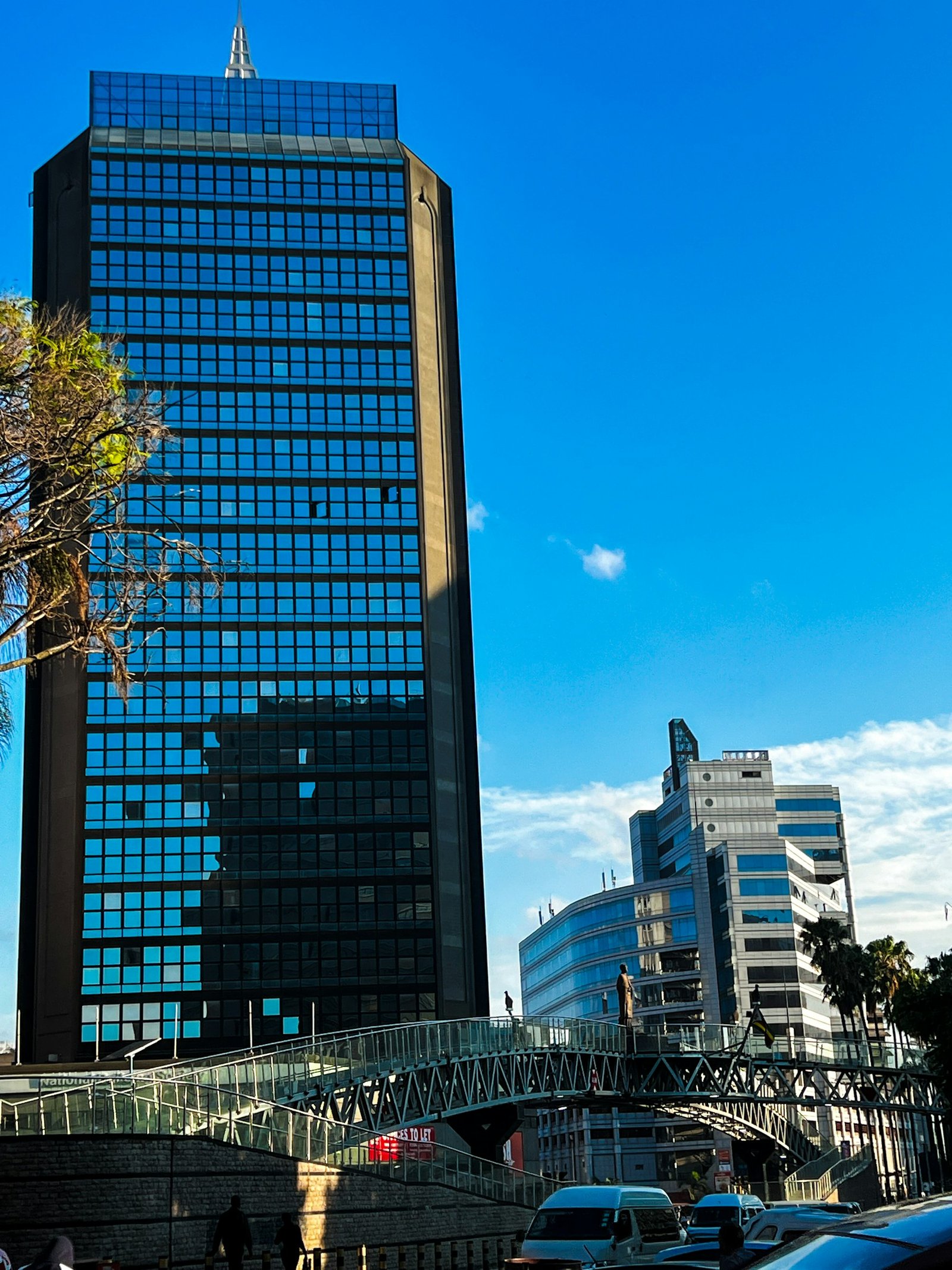 Glass office towers in Harare's central business district, a steel pedestrian footbridge crossing the street below